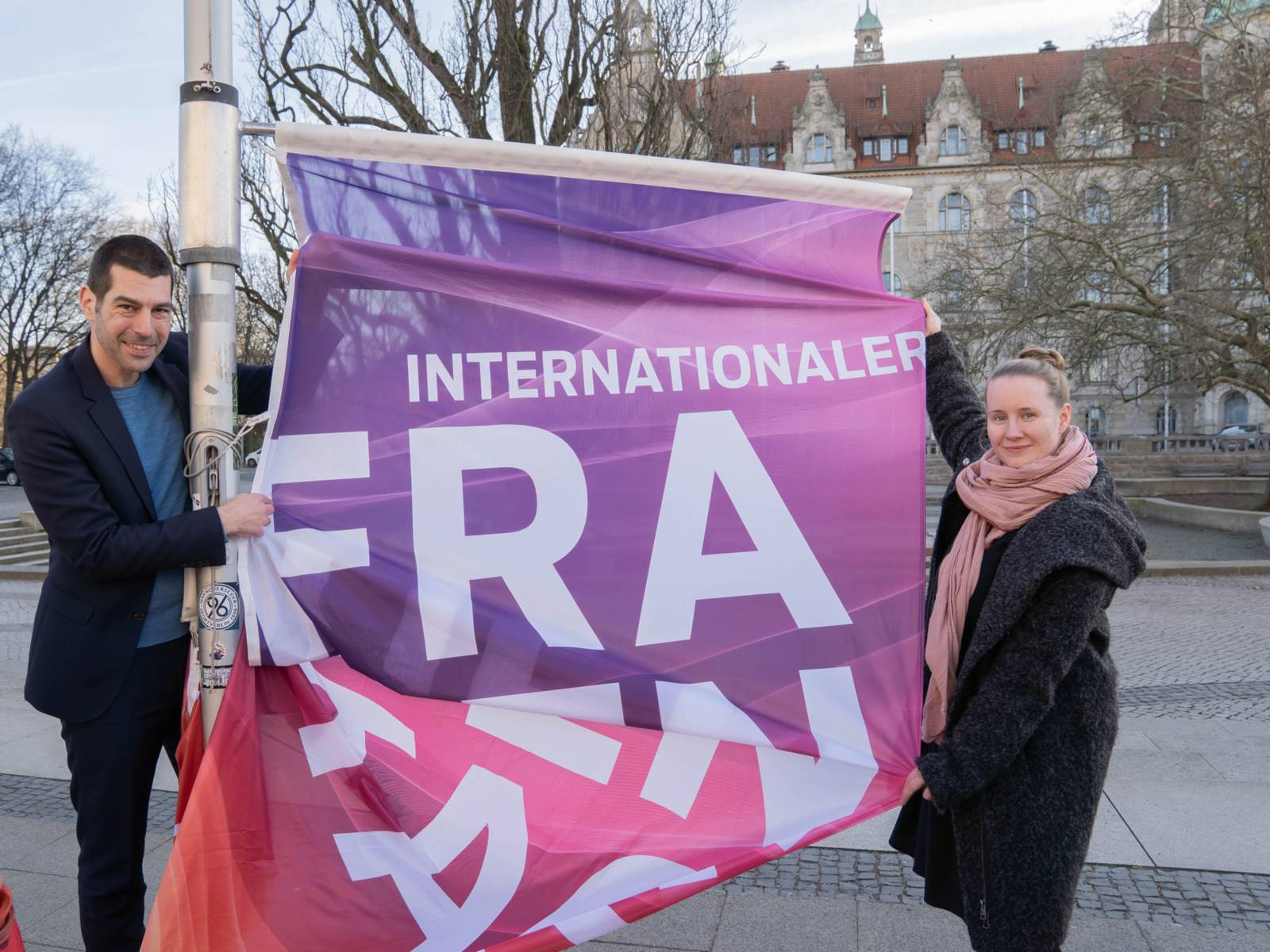 Ein Mann und eine Frau halten eine Flagge an einem Masten in den Händen. Die Flagge in den Farben Lila bis Pink ist zu lesen Internationaler FRA (weiterer Text liegt in Falten)