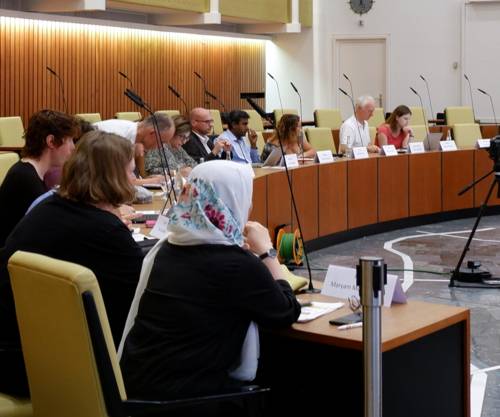 12 Personen sitzen in einem Halbkreis an holzvertäfelten Tischen. Von den Tischen ragen Mikrofone auf. Die Personen befinden sich im Ratssaal der Landeshauptstadt Hannover.