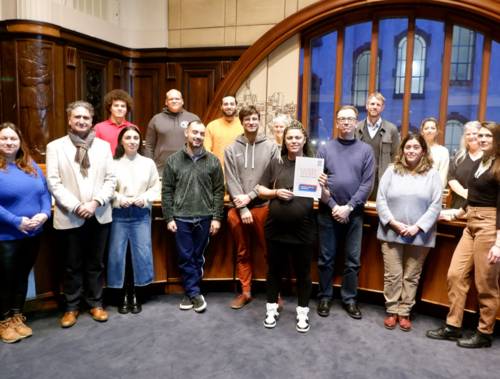 16 Personen stehen für ein Gruppenfoto vor einer halbrunden Plenar-Tisch-Reihe. Sie lächeln in die Kamera. Eine Frau in der Bildmitte hält den WIR2.0-Maßnahmenkatalog in die Kamera. Im Hintergrund ein großes Fenster in einen Innenhof des Neuen Rathauses.