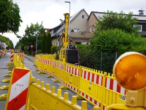 Rot-weiß gestreifte Markierungen und gelbe Absperrungen mit einer gelben Lamp links, dahinter eine Straße und Wohnhäuser.