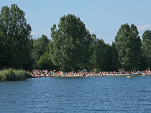 blaues Wasser, im Hintergrund badende Menschen an der Uferzone des Altwarmbüchener Sees