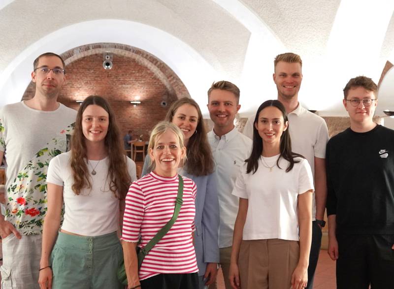 Eine Gruppe von fünf Personen schaut in die Kamera. Dahinter befindet sich ein großer Raum mit einem weißen Gewölbebögen an der Decke.