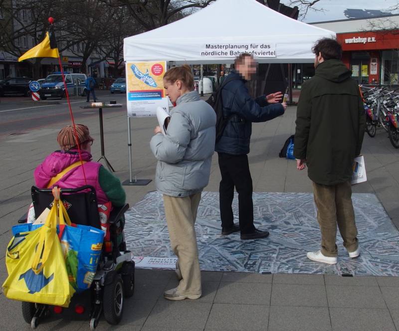 Auf dem Bild sind vier Personen zu sehen. Ganz links sitzt eine Person in einem elektrischen Rollstuhl und unterhält sich mit einer rechts daneben stehenden Person, die einen Notizblock in der Hand hält. Etwas im Hintergrund stehen zwei weitere Menschen, das Gesicht des einen ist unkenntlich gemacht. Auch sie unterhalten sich miteinander. Weiter im Hintergrund ist ein Pavillon zu sehen, der die Aufschrift "Masterplanung für das nördliche Bahnhofsviertel" trägt.