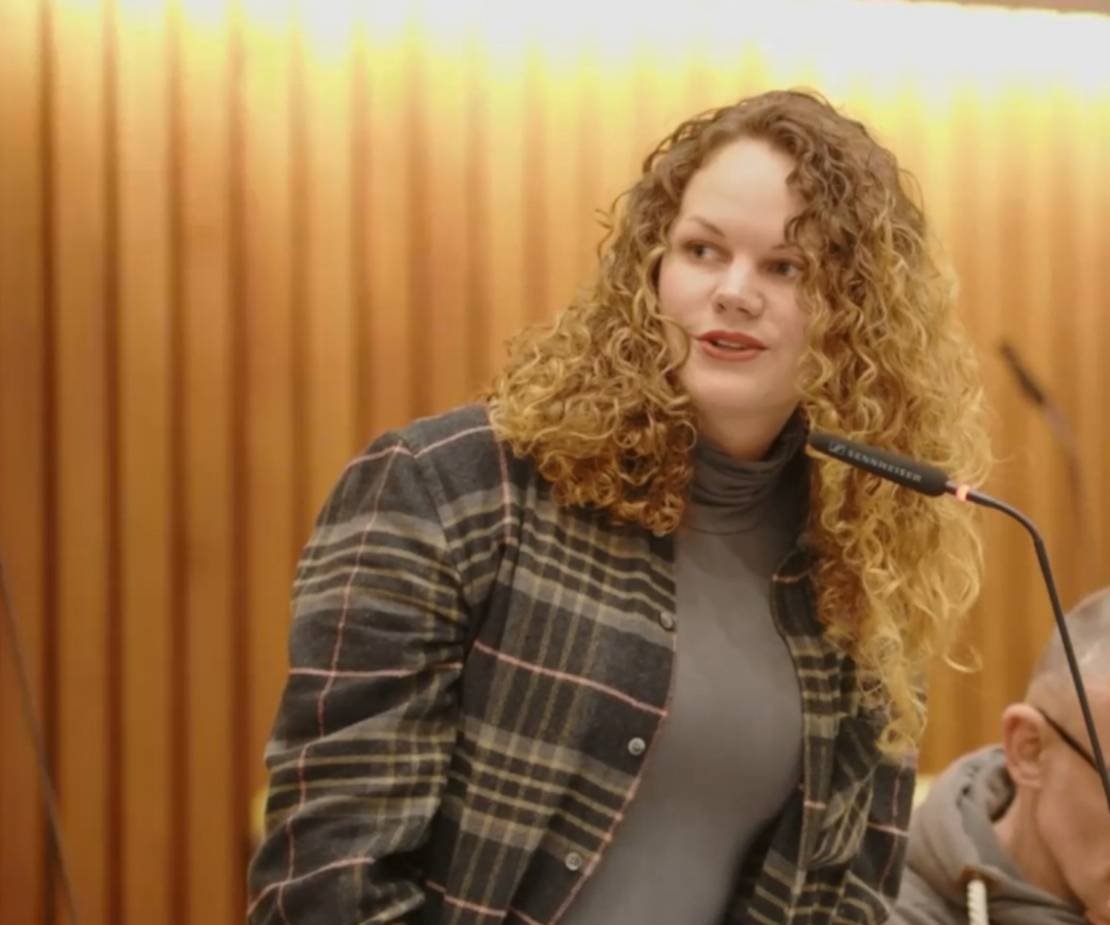 Eine Frau mit langen blonden Locken spricht in ein Mikrofon. Sie trägt ein kariertes Hemd.