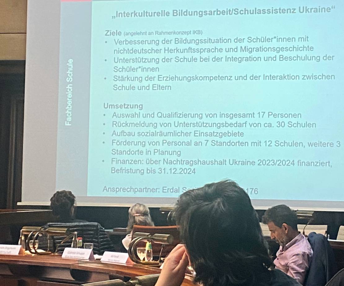 Eine Folie auf einer Leinwand nimmt fast das gesamt Bild ein. Überschrift der Folie ist "Interkulturelle bildungsarbeit/Schulassistenz Ukraine". Im Vordergrund die Köpfe von vier Personen. Drei davon schauen zu der Leinwand.