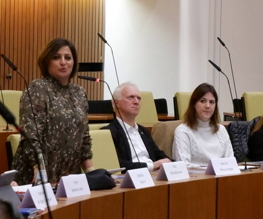 Eine Frau mit dunklen Haaren steht an einem Tisch und spricht in ein Mikrofon. Neben ihr sitzt ein Mann im schwarzen Sakko. Sein Gesicht drückt keine Zustimmung aus.