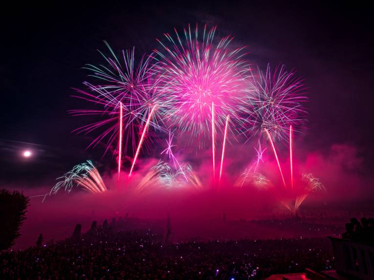 Feuerwerk in Pink und Blau erleuchtet den Nachthimmel über einer großen Menschenmenge. Dichte Rauchschwaden im Vordergrund.