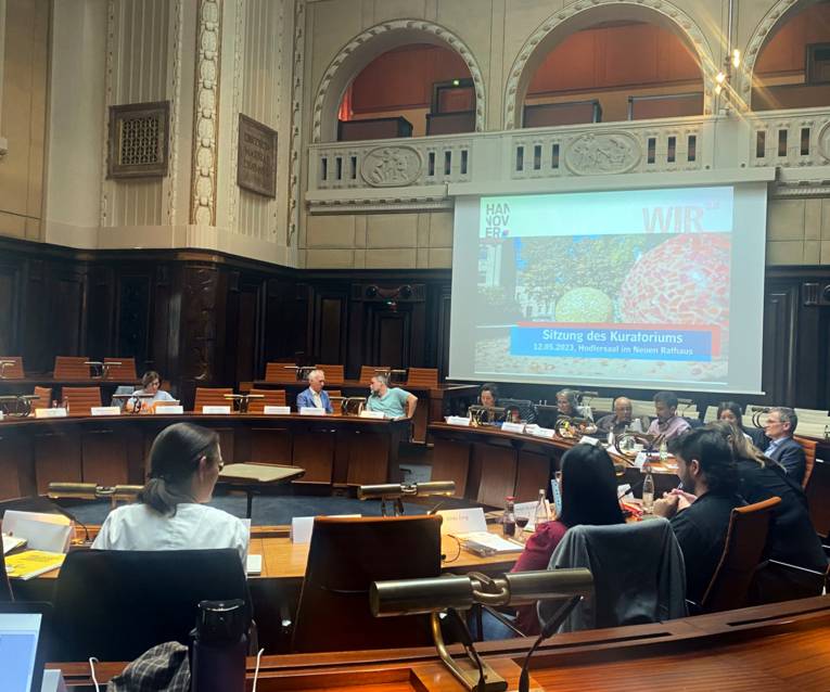 Eine Sitzung im Hodlersaal im Neuen Rathaus. Der Saal ist an den Wänden mit dunklem Holz vertäfelt. Die fest installierten Holztische sind in zwei Reihen kreisrund angeordnet. An den Tischen sitzen über das Bild verteilt 13 Personen, die teilweise mit ihren NachbarInnen Diskutieren. Rechts im Hintegrund ist auf einer Leinwand eine Folie mit dem Text "WIR2.0 Sitzung des Kuratoriums" zu sehen.