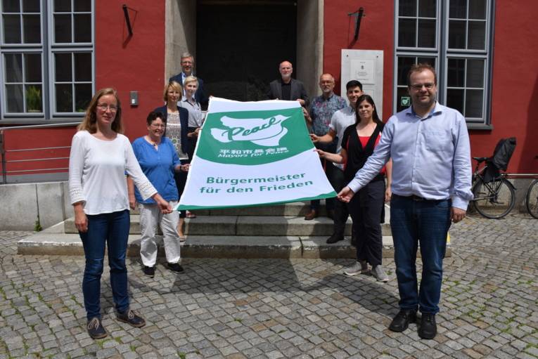 10 Menschen halten die Mayors for Peace Flagge in Greifswald