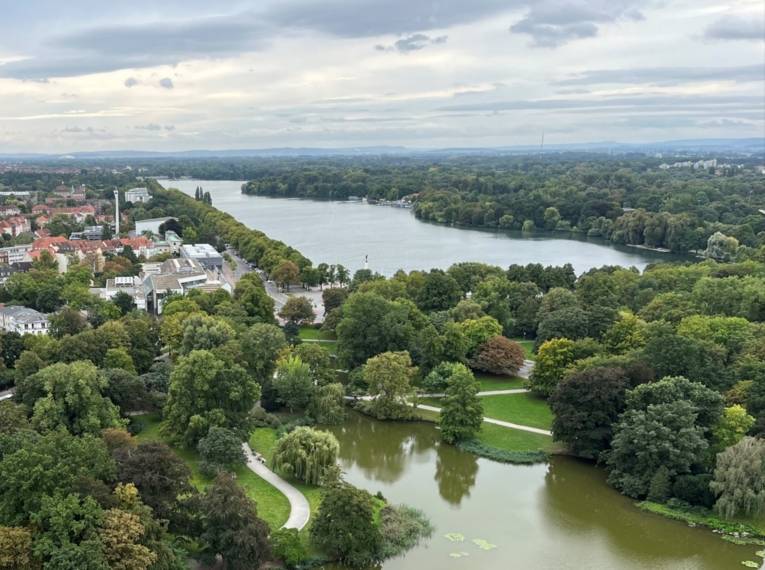 Blick von oben auf einen großen See, umringt von grünen Bäumen, links auch Häuser.